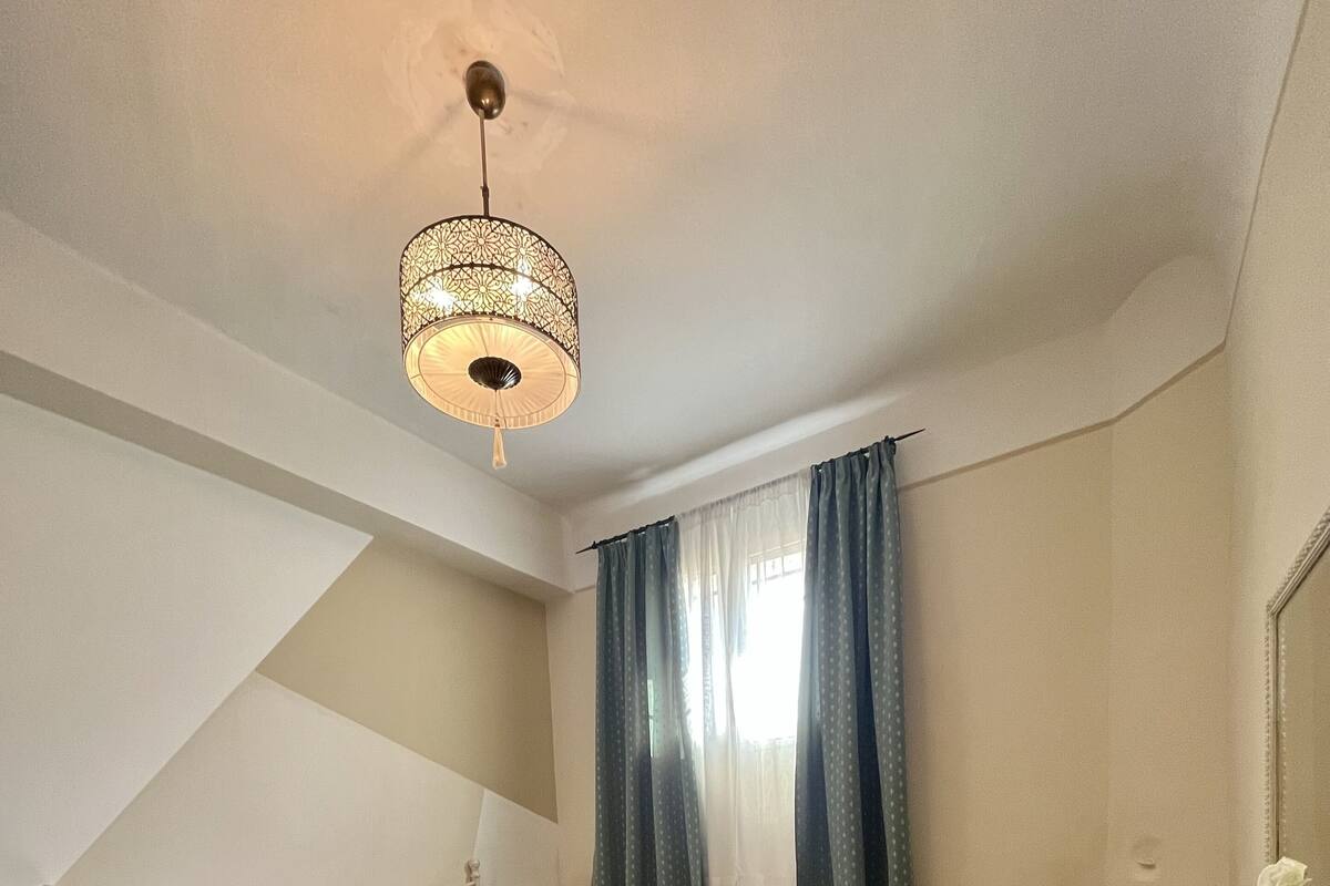A bedroom with a bed, a bedside table, a lamp, a mirror, a fireplace, and a window with curtains.