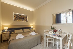 Una habitación de hotel con una cama, mesitas de noche, un escritorio con sillas, un espejo y un cuadro en la pared.