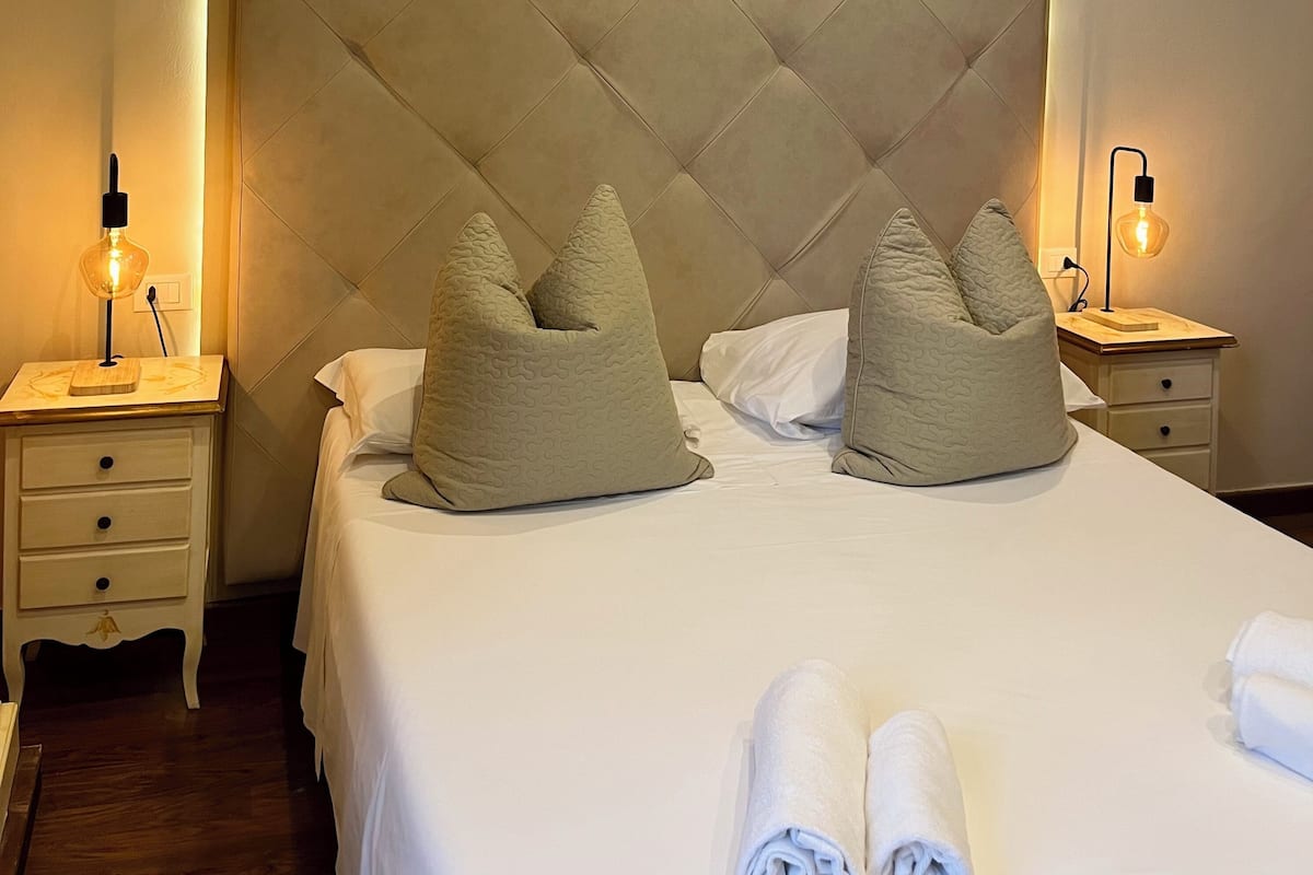 A neatly made bed with pillows, a bedside table with a lamp, and a wall-mounted light.