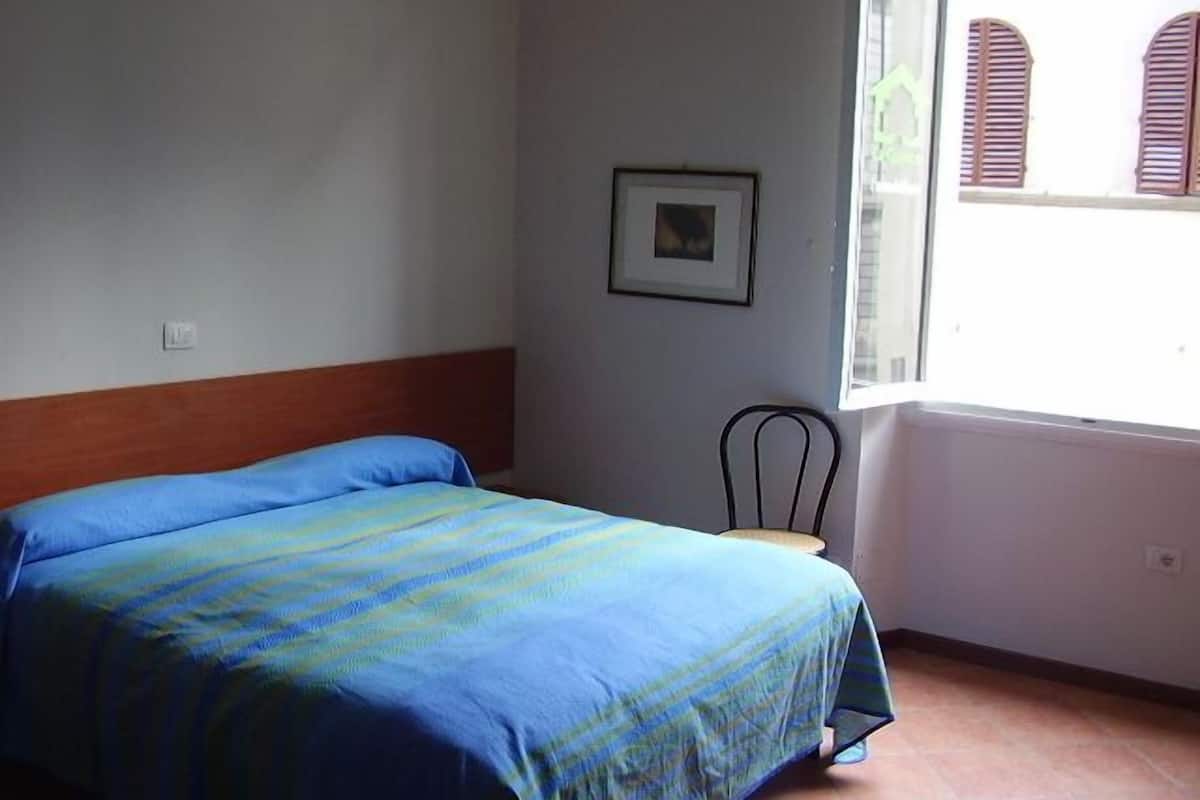 Een slaapkamer met een bed, een stoel, een raam en een foto aan de muur.