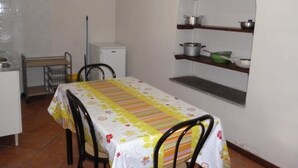 Een klein eetgedeelte met een tafel bedekt met een bloemenkleed, twee stoelen, een witte koelkast, en een plank met keukengerei.