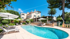 A swimming pool with lounge chairs and umbrellas, surrounded by a landscaped garden and a large building with multiple windows. - Mas Djoliba (Antibes)
