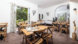 A dining area with wooden tables and chairs, white walls, and a view of greenery outside. - Mas Djoliba (Antibes)