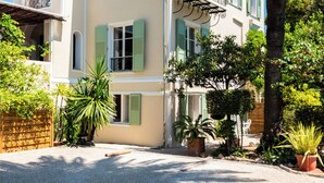 A two-story building with a beige facade, green shutters, and a red-tiled roof. There are potted plants and a palm tree in the front yard. - Mas Djoliba (Antibes)