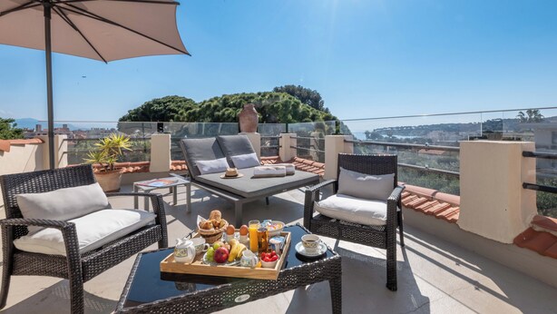A balcony with wicker furniture, a tray of breakfast, and a view of a cityscape and greenery. - Mas Djoliba (Antibes)