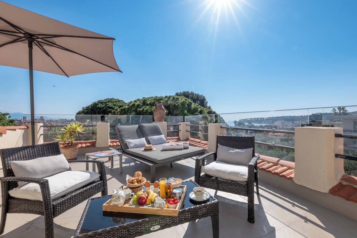 A balcony with wicker furniture, a tray of breakfast, and a view of a cityscape and greenery.