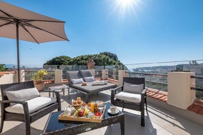 A balcony with wicker furniture, a tray of breakfast, and a view of a cityscape and greenery. - Mas Djoliba (Antibes)