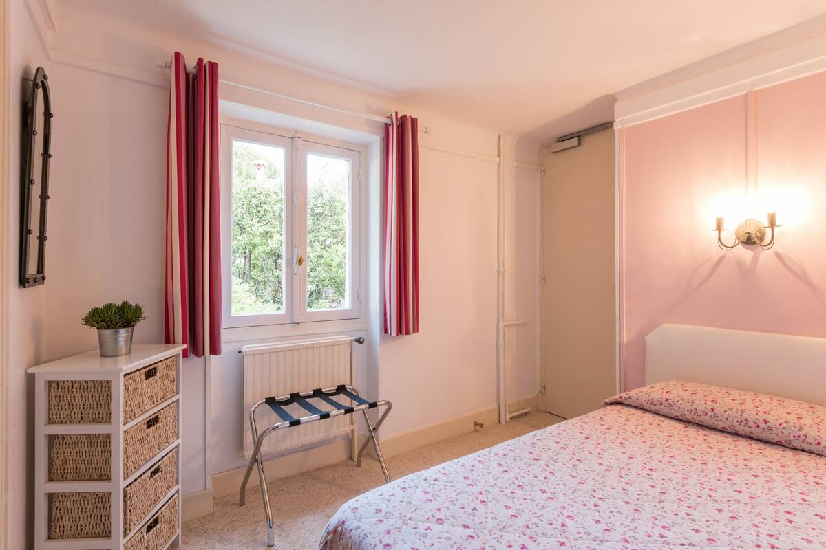 A bedroom with a bed, a radiator, a window with curtains, a bedside table with a plant, and a wall-mounted light fixture.