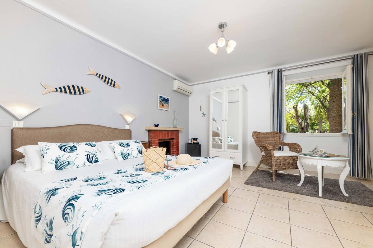 A bedroom with a bed, a chair, a small table, a fireplace, and a window with a view of trees.