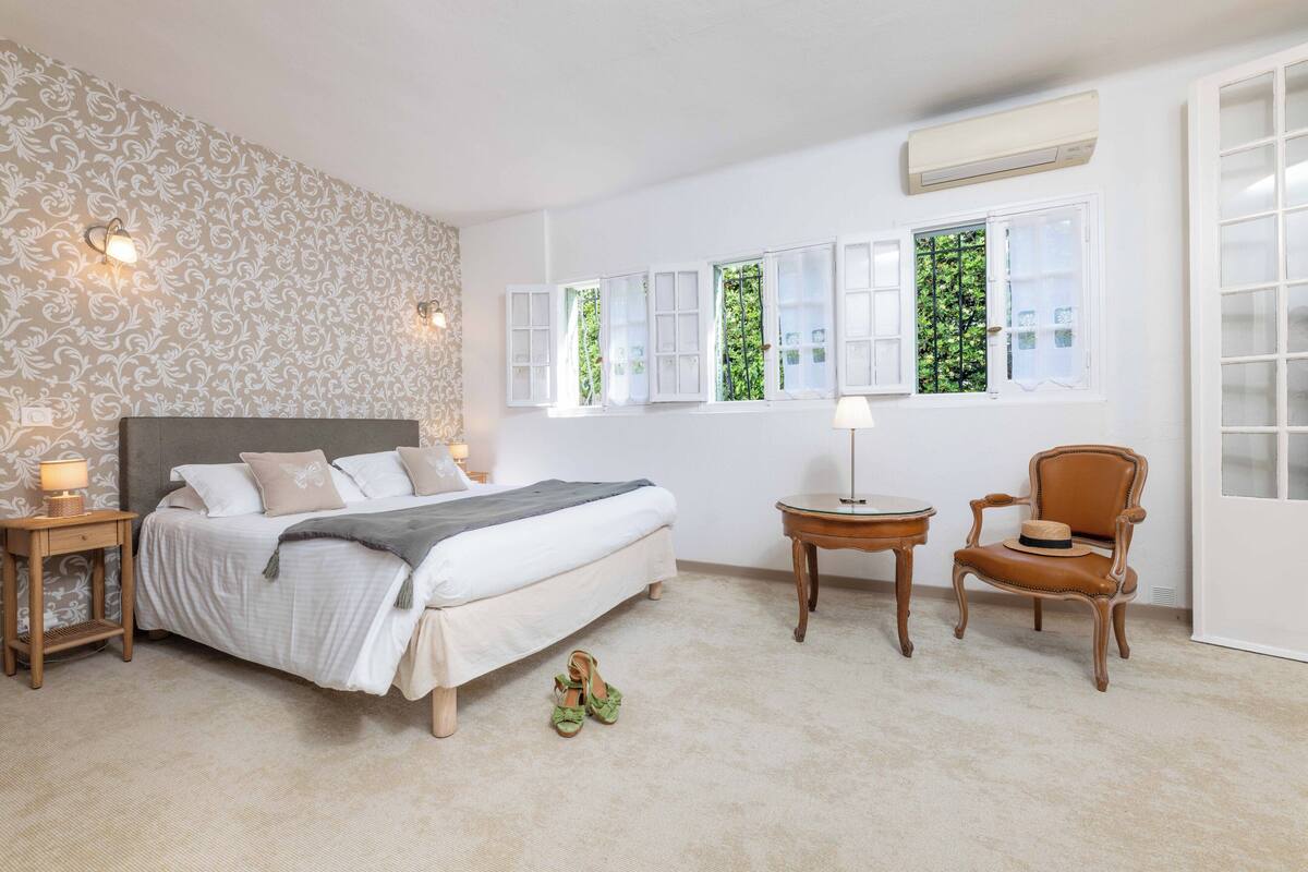 A bedroom with a bed, bedside tables, a chair, and windows with shutters.