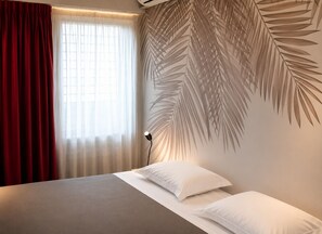 A hotel room with a bed, a wall-mounted air conditioner, a window with sheer curtains, and a wall with a palm leaf pattern. - Hotel Alnea (Cannes)