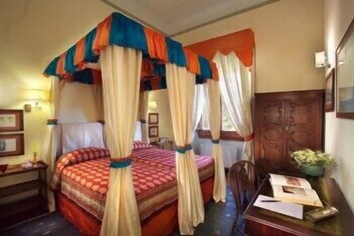Una cama con dosel con un edredón estampado en rojo y naranja, una mesita de noche de madera con una lámpara, y un escritorio con papeles y una planta.