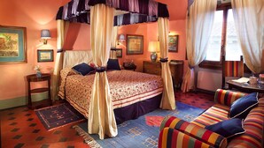 Una cama con dosel y cortinas, un edredón floral, un sofá a rayas y una alfombra estampada en una habitación cálidamente iluminada con paredes anaranjadas.