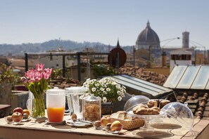 Una mesa de desayuno con comida y bebidas en una azotea con vistas a un paisaje urbano de edificios históricos.