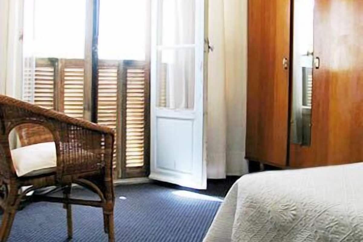 Quarto de hotel com uma cadeira de vime, uma cama com colcha branca e um guarda-roupa de madeira.
