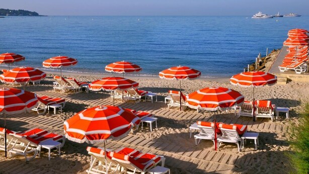 A beach with rows of white lounge chairs and red and white striped umbrellas. - Hotel Pierre Loti (Antibes)