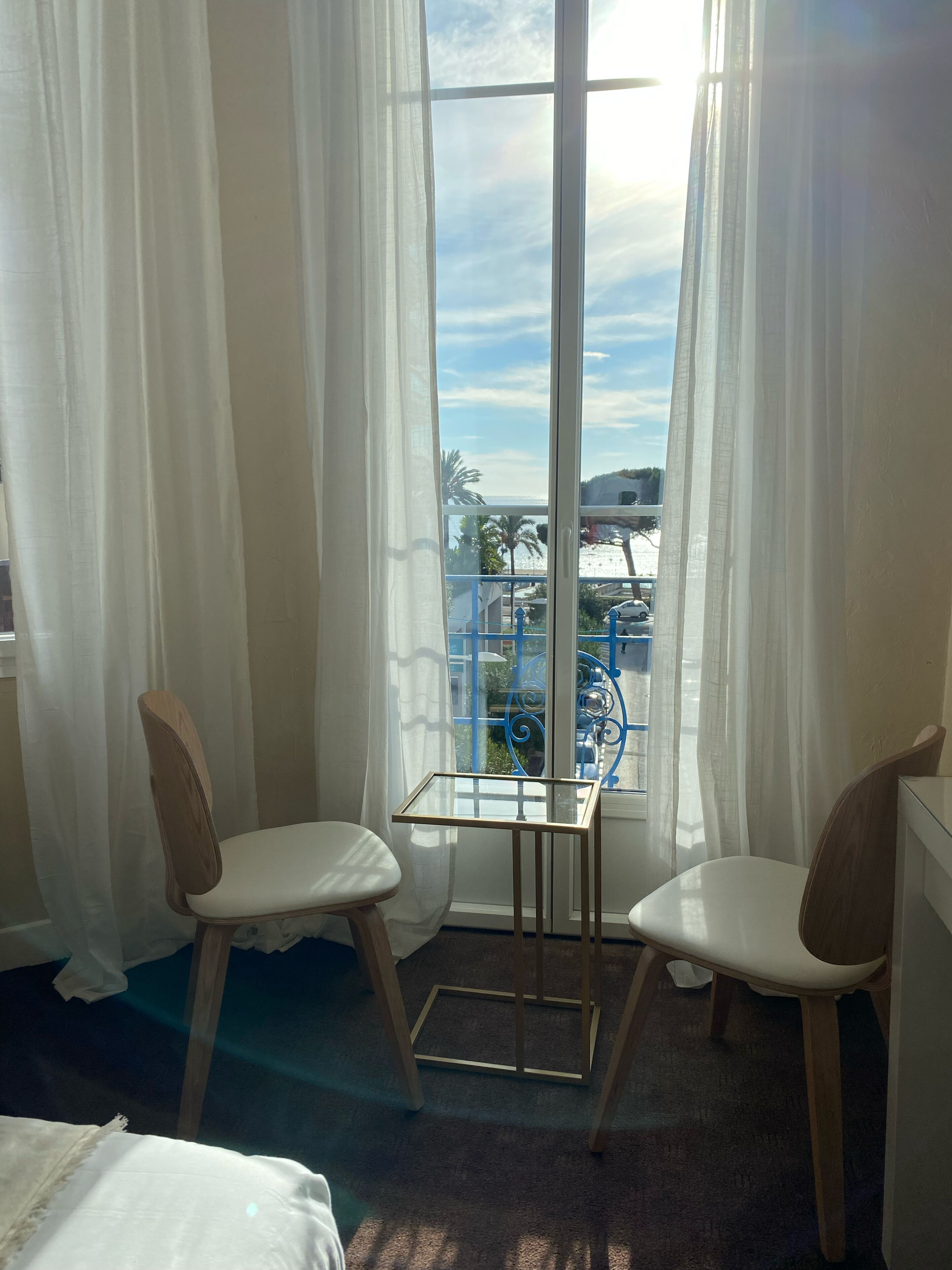 Une pièce avec deux chaises en bois, une petite table en verre et une vue sur une balustrade bleue et des palmiers, le tout visible à travers de fines rideaux.