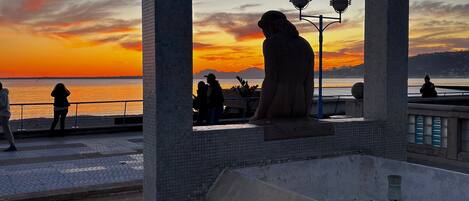 Vista al atardecer enmarcada por un gran arco de piedra con vista a un paseo marítimo donde hay gente sentada y caminando.