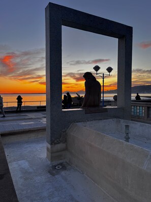 A sunset view framed by a large stone archway overlooking a waterfront with people sitting and walking. - Hôtel Eden - La Baigneuse (Antibes)