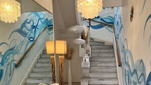 Una escalera con acabado de mármol, un sofá blanco con cojines, un tapiz tejido y obras de arte mural con diseños abstractos en azul y blanco.