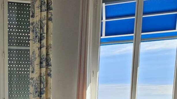 Un dormitorio con cama, dos sillas, una mesita con un jarrón de flores y un ventanal con vistas al mar.
