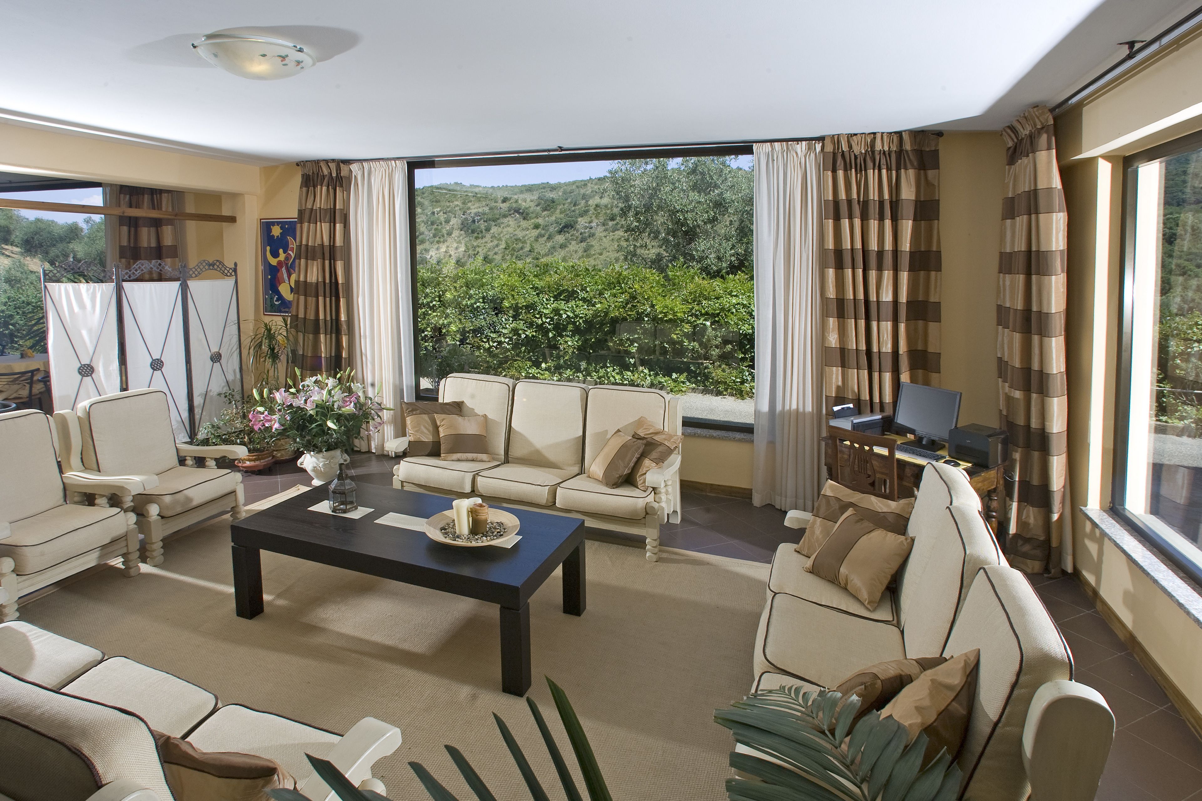 A spacious living room with a sofa, armchairs, and a coffee table. There is a large window with curtains, a television, and a plant.