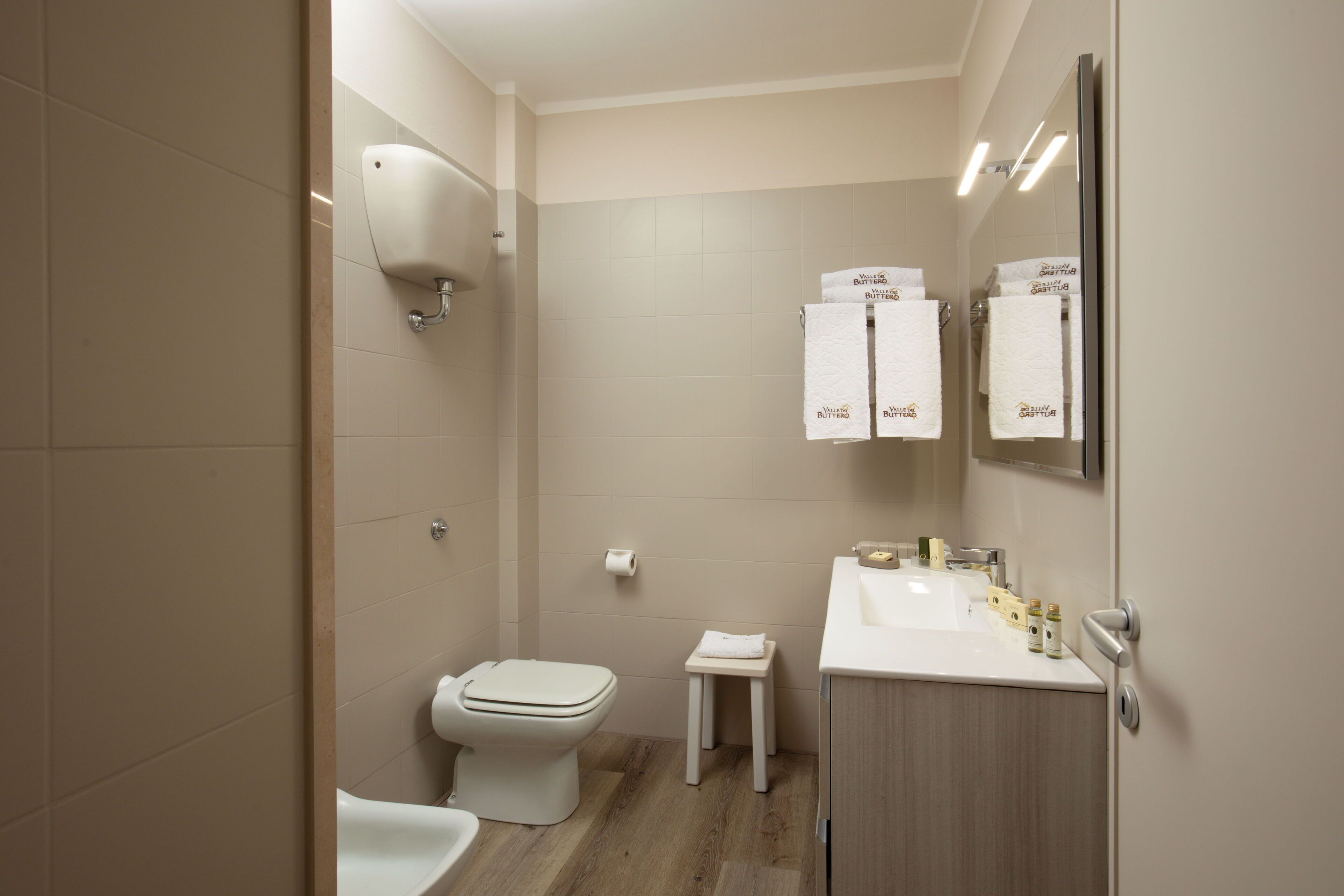 A modern bathroom with a toilet, sink, and a small stool. There are towels hanging on the wall and a mirror above the sink.