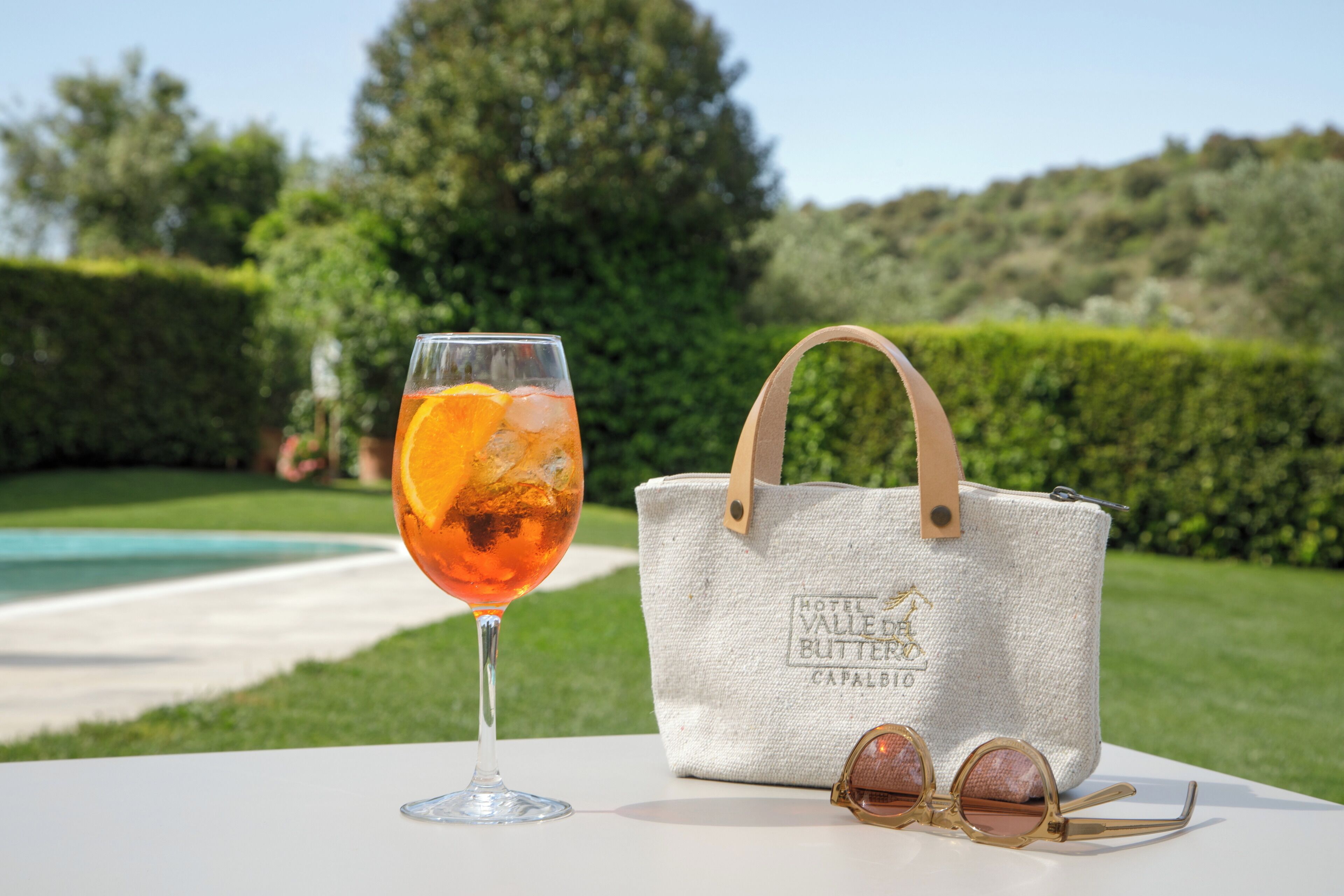 A glass of orange juice, a white tote bag, and sunglasses on a table with a pool and greenery in the background.