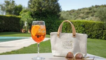 A glass of orange juice, a white tote bag, and sunglasses on a table with a pool and greenery in the background.