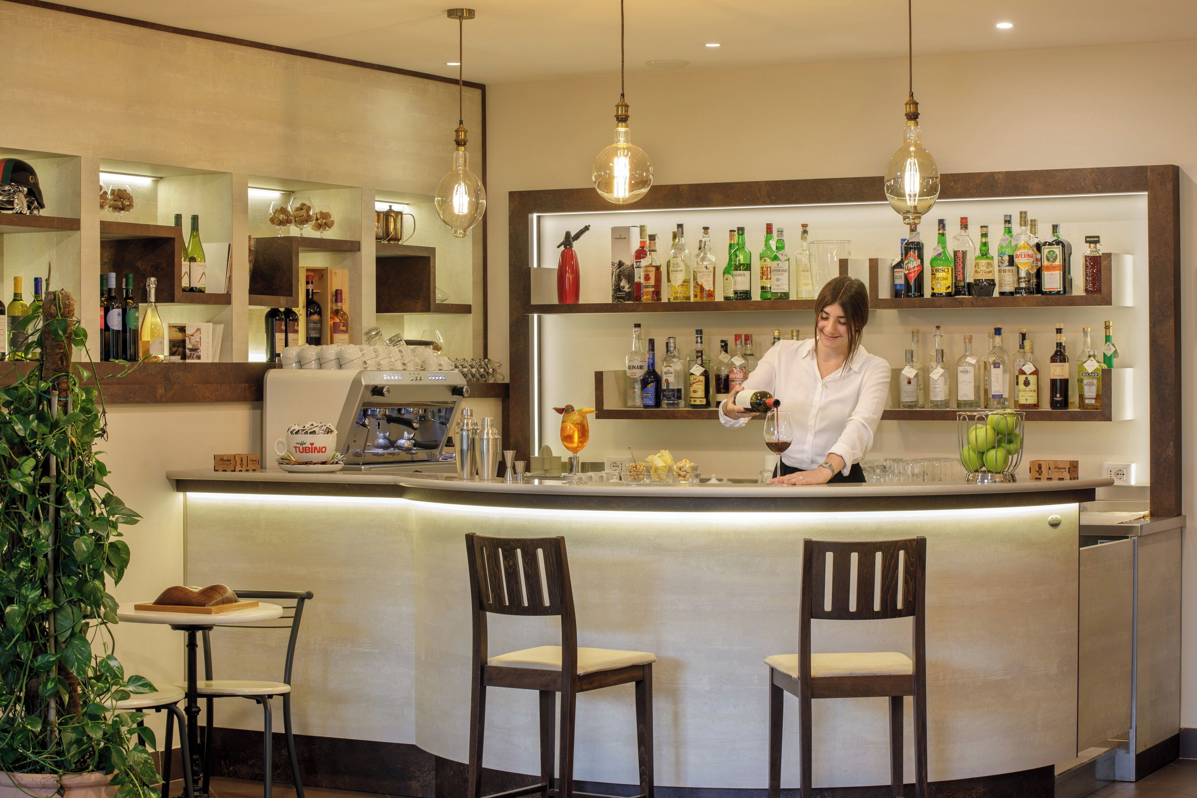 Zona de bar con un cantinero, estantes llenos de botellas de licores y dos taburetes.