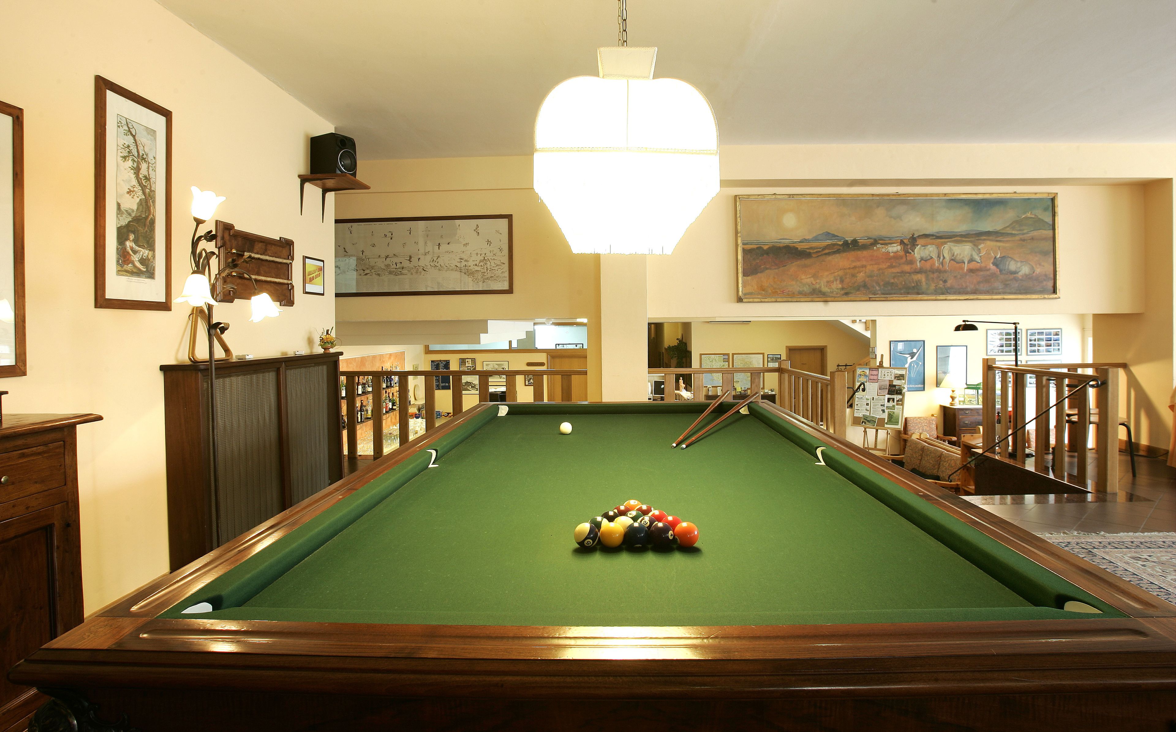 Une table de billard dans une pièce avec un escalier, des œuvres d’art encadrées et un luminaire suspendu.