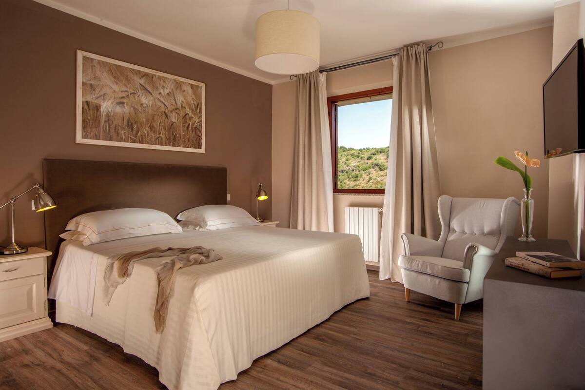 Ein Schlafzimmer mit einem großen Bett, einem Sessel, einem Schreibtisch und einem Fenster mit Vorhängen.