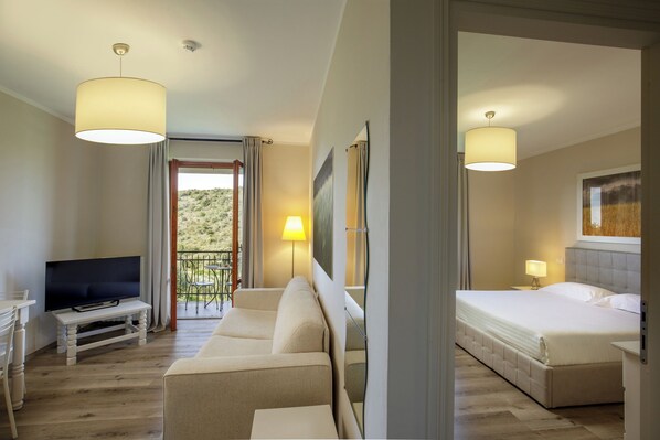 Habitación de hotel con sofá, cama, televisor en un mueble bajo y balcón con vista a zonas verdes.