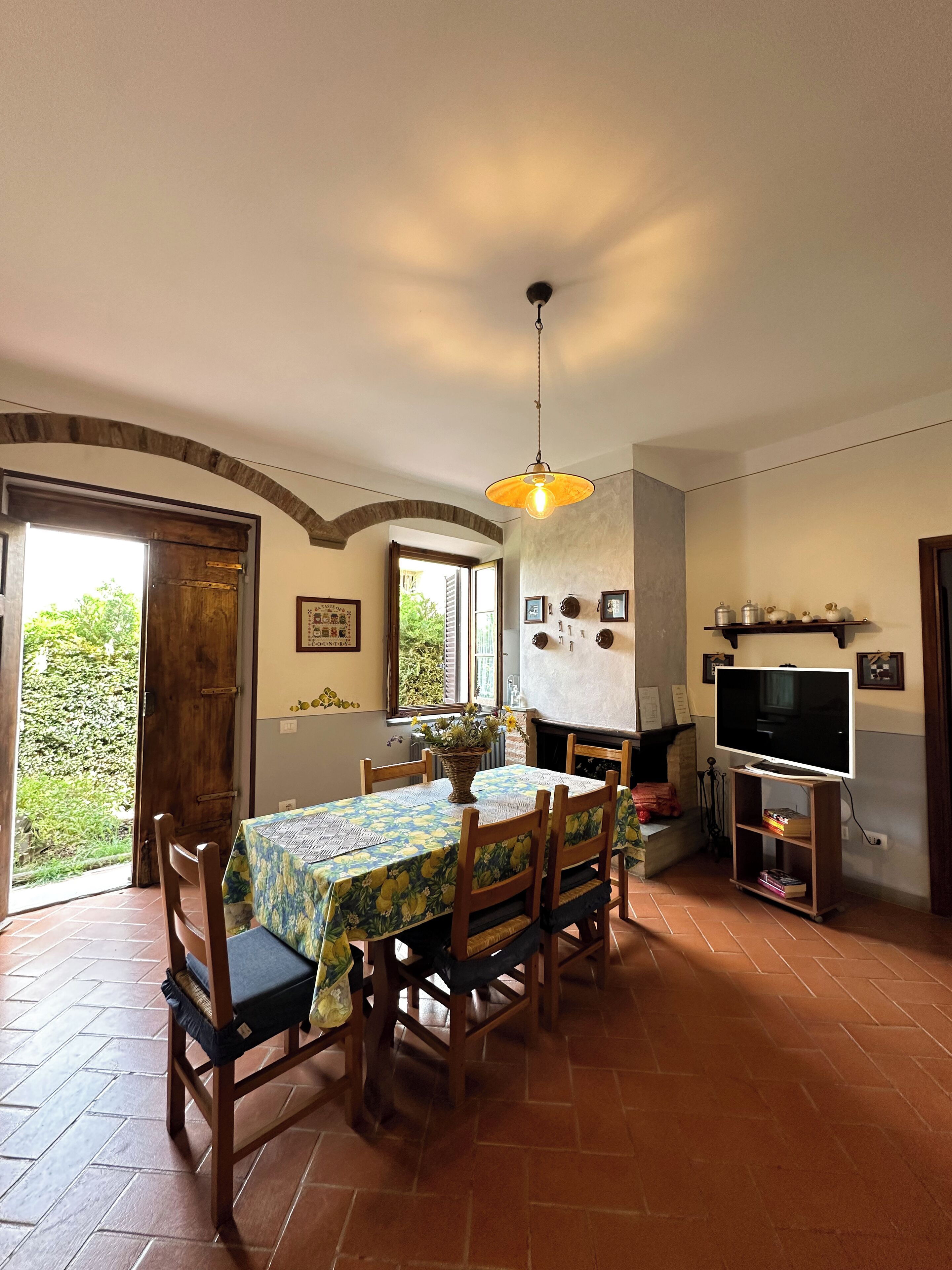 A dining area with a table set for six, surrounded by wooden chairs. A hanging light fixture is above the table, and a television is mounted on the wall to the right.