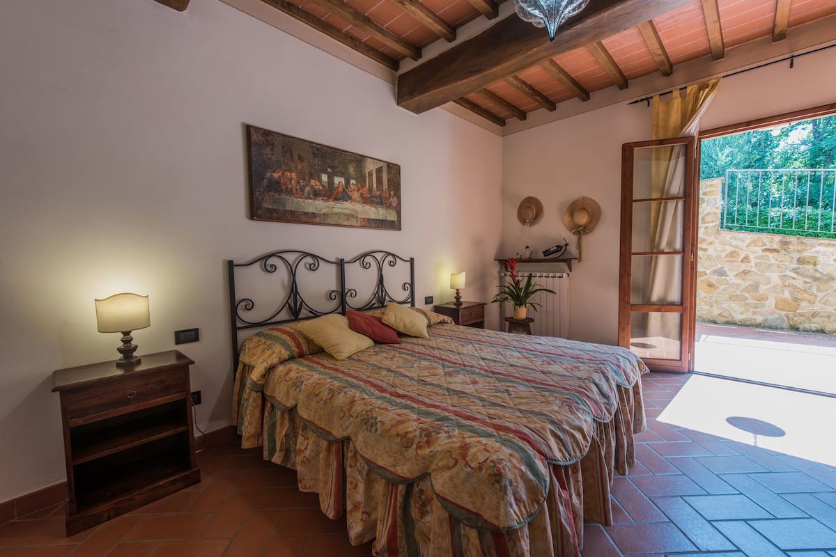 Un dormitorio con cama, mesita de noche, lámpara, un cuadro y vista al exterior.