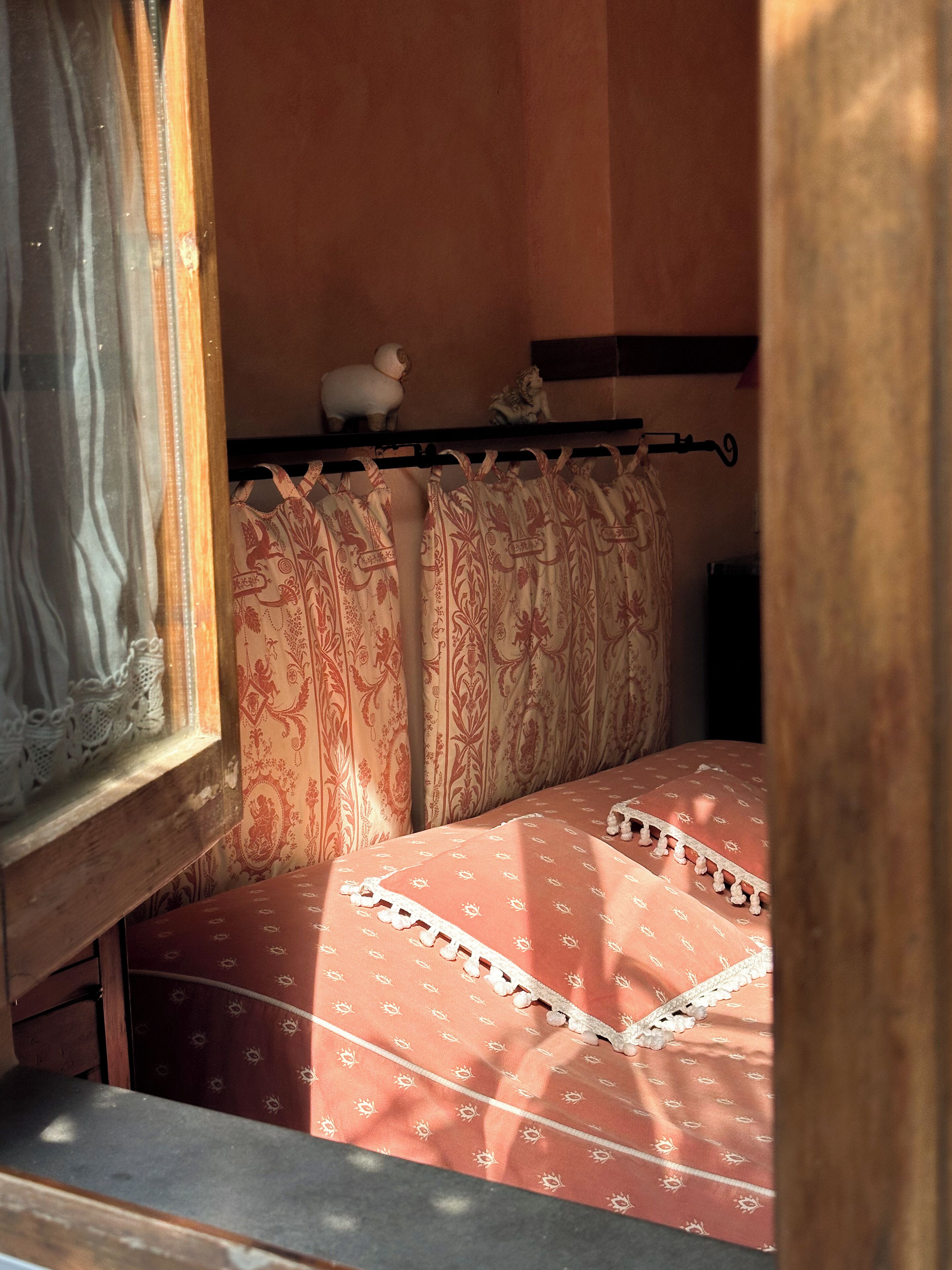A bedroom with a bed covered in a patterned bedspread, a window with lace curtains, and a shelf with decorative items.