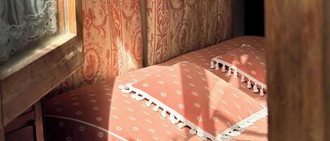 A bedroom with a bed covered in a patterned bedspread, a window with lace curtains, and a shelf with decorative items.