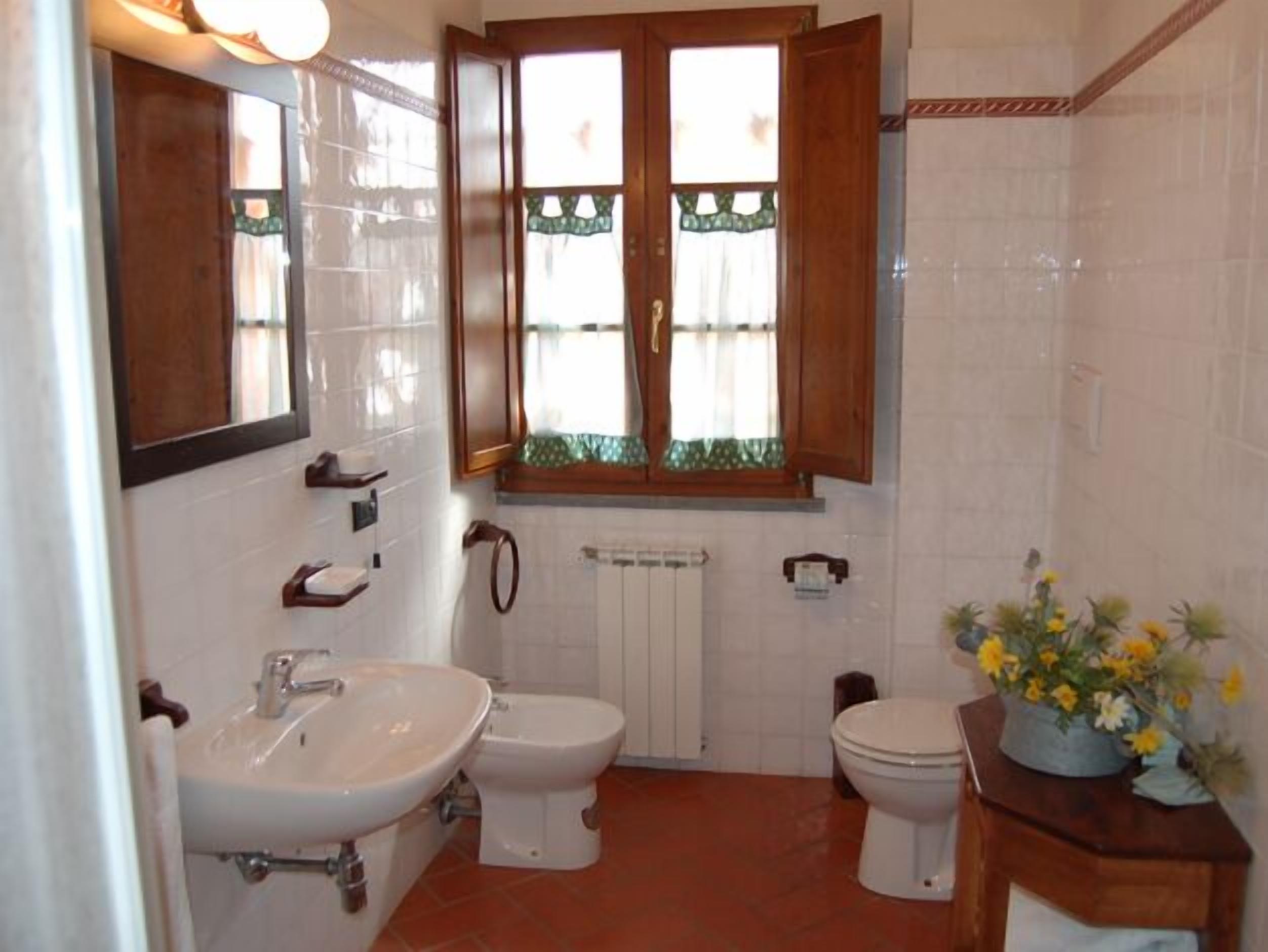 Une salle de bain équipée d’un lavabo, de toilettes et d’un bidet. Il y a une fenêtre avec des rideaux et un meuble en bois au-dessus des toilettes.