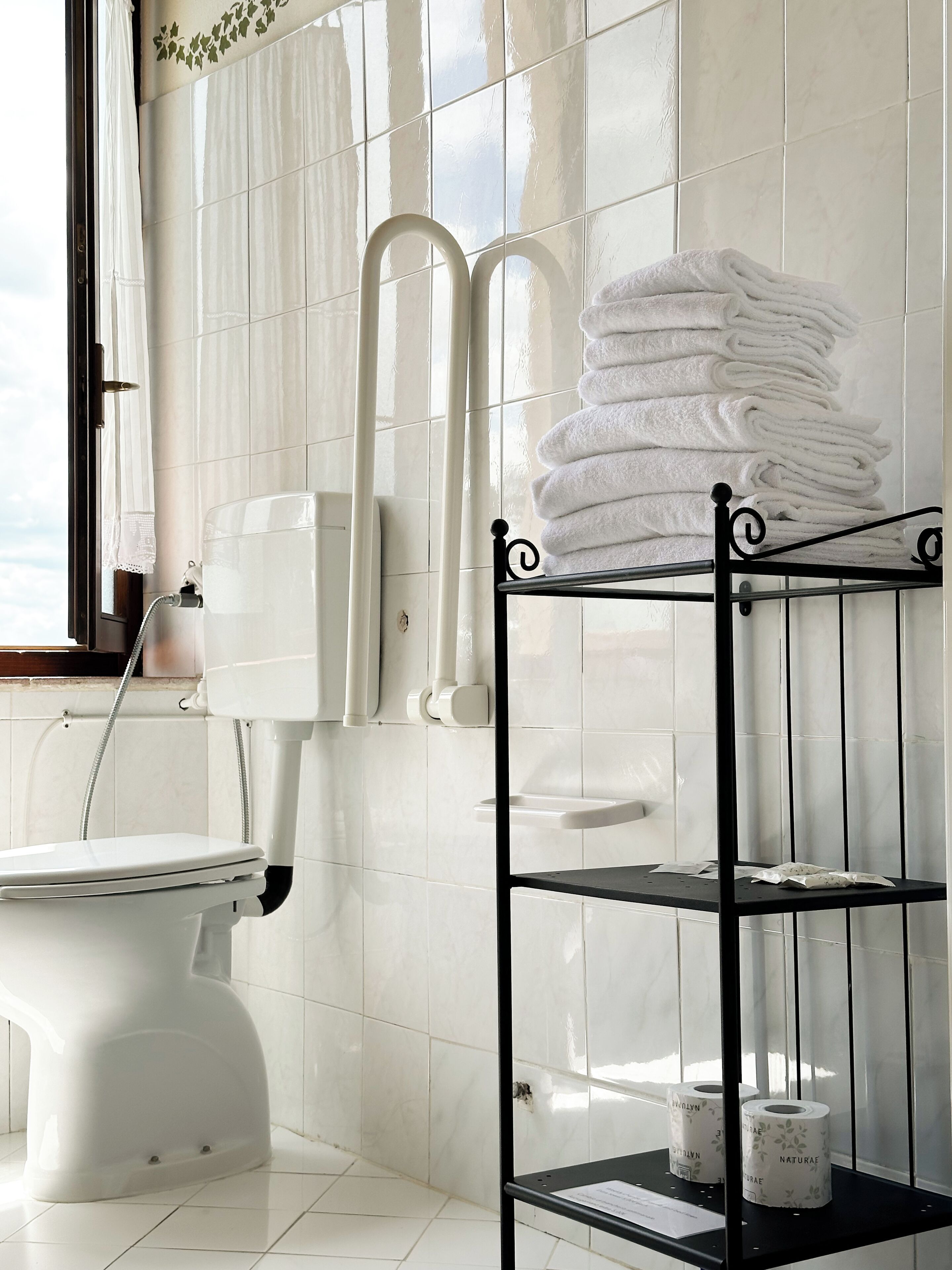 A bathroom with a toilet, a towel rack with folded towels, a shelf with toilet paper, and a window with curtains.