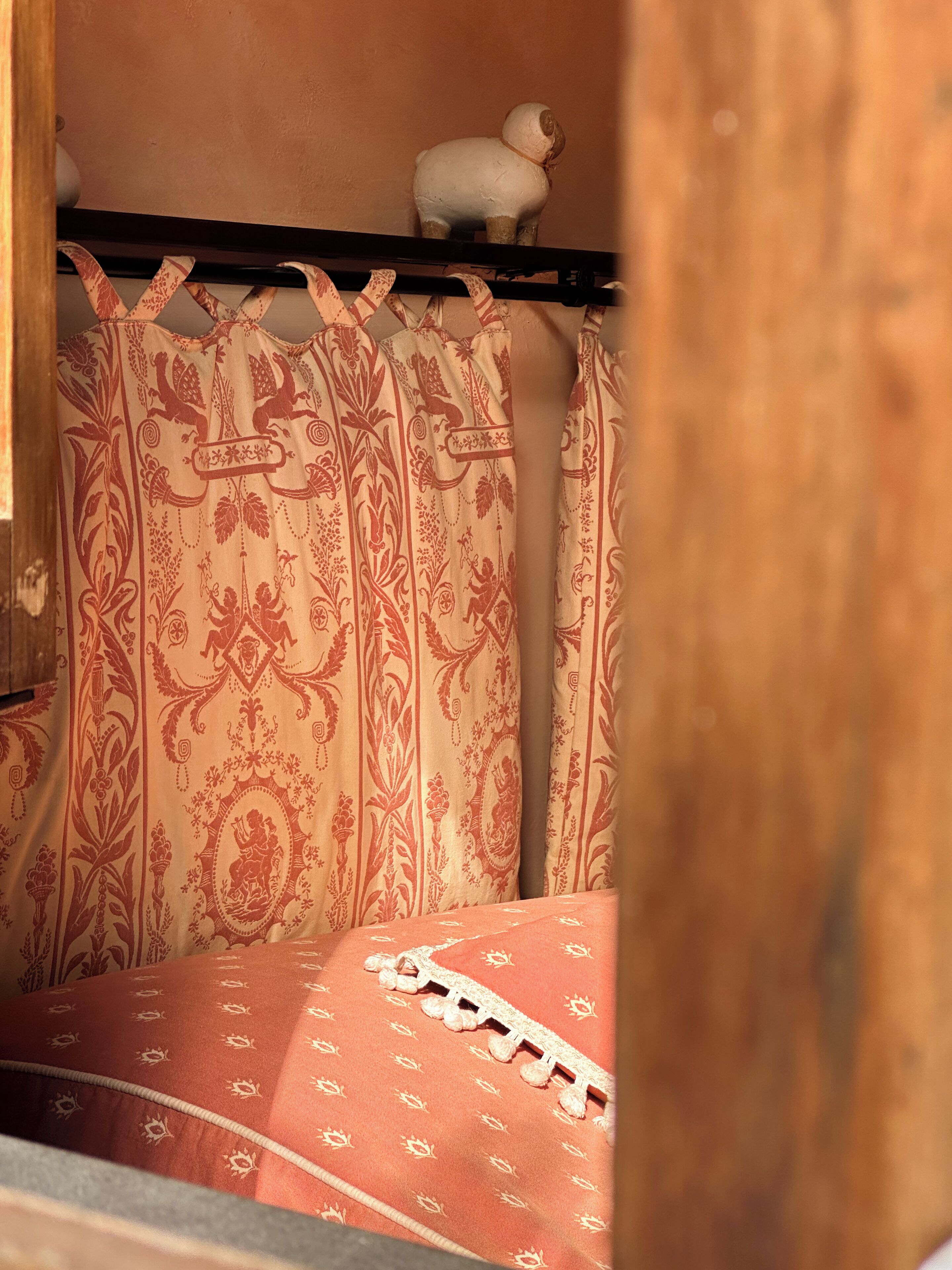 A room with a patterned curtain, a bed with a patterned bedspread, and a small white figurine on a shelf.