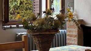 Une pièce avec une table mise pour le repas, un panier en osier rempli de fleurs, un radiateur et une fenêtre dont les volets sont ouverts.