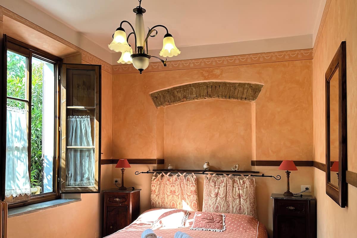 Un dormitorio con cama, dos mesitas de noche, una araña y una ventana con cortinas, con vistas a zonas verdes.
