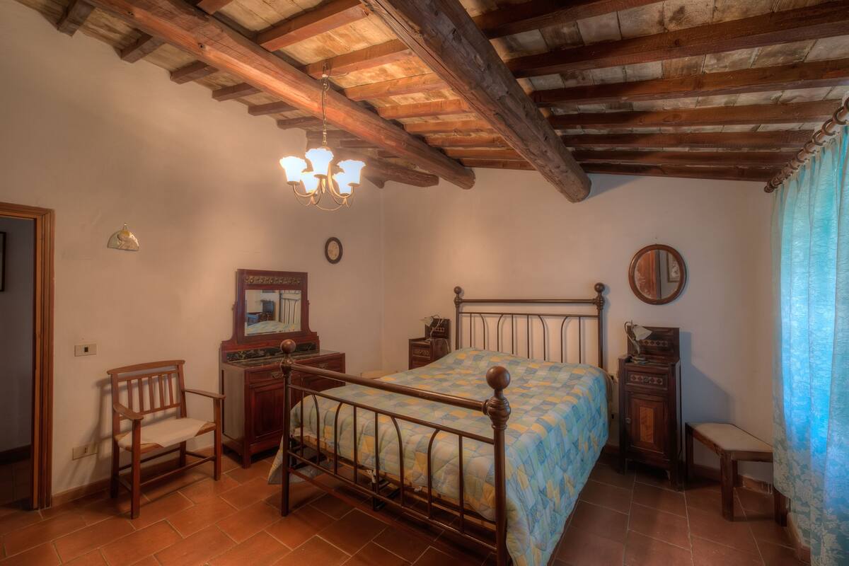 Un dormitorio con estructura de cama de madera, candelabro, silla de madera, una mesita y un espejo en la pared.