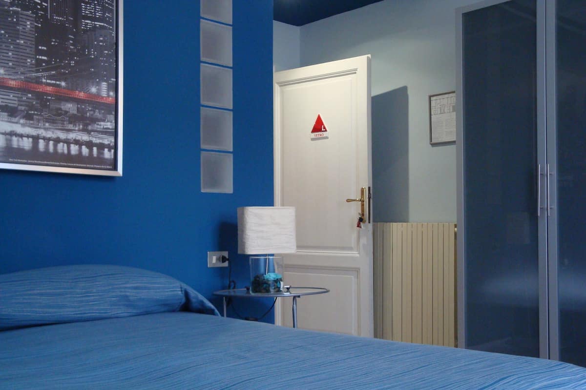 A hotel room with a bed, a nightstand, a lamp, a door with a red triangle sign, and a window with a cityscape view.