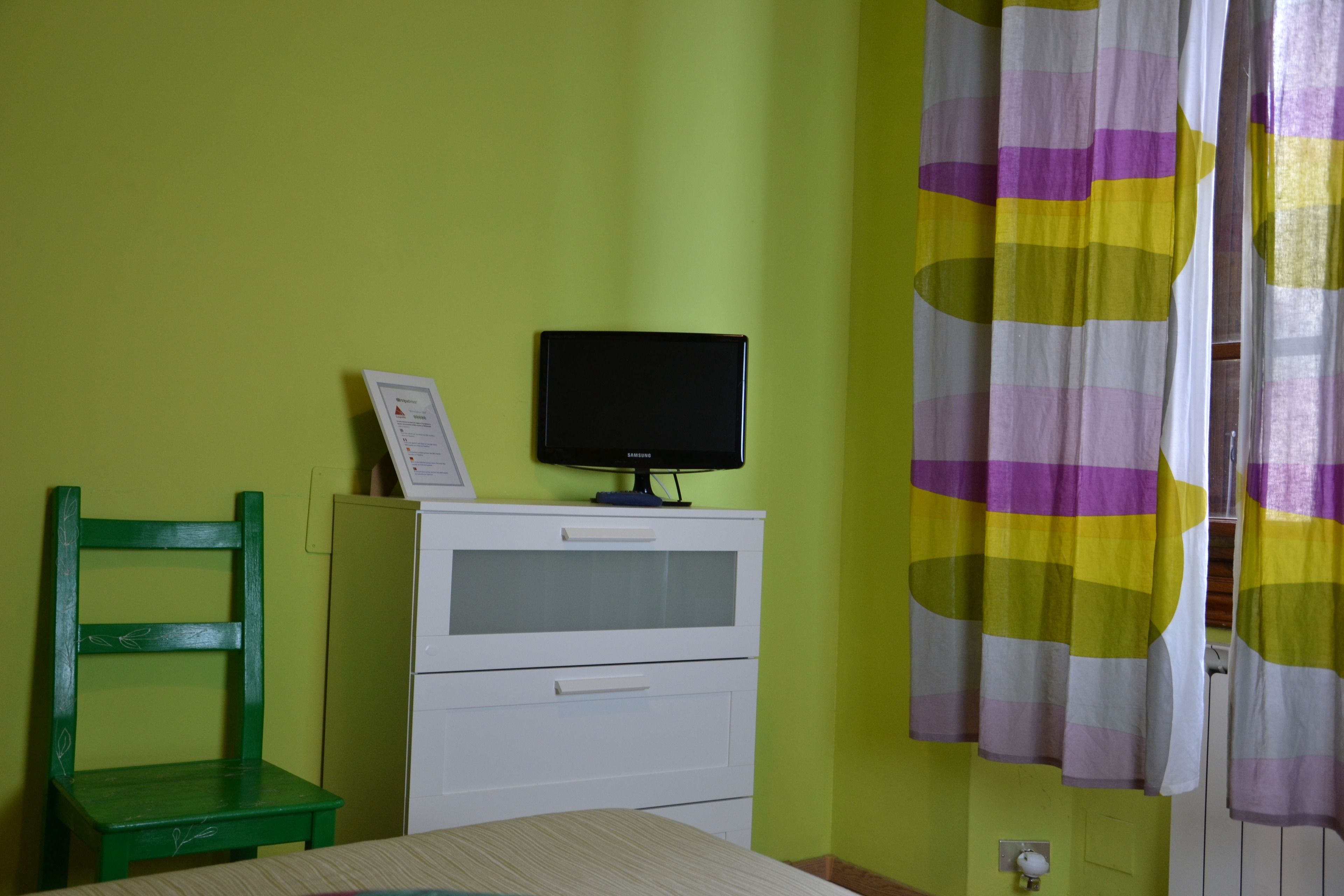 Ein Zimmer mit grüner Wand, einem weißen Schrank mit Fernseher darauf, einem grünen Sessel und einem gestreiften Vorhang.
