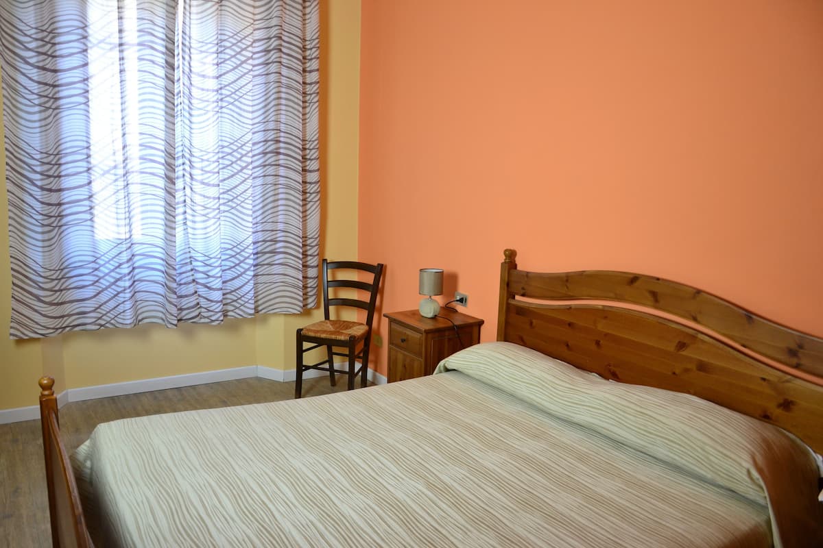 A bedroom with a bed, a chair, a bedside table, and a window with curtains.