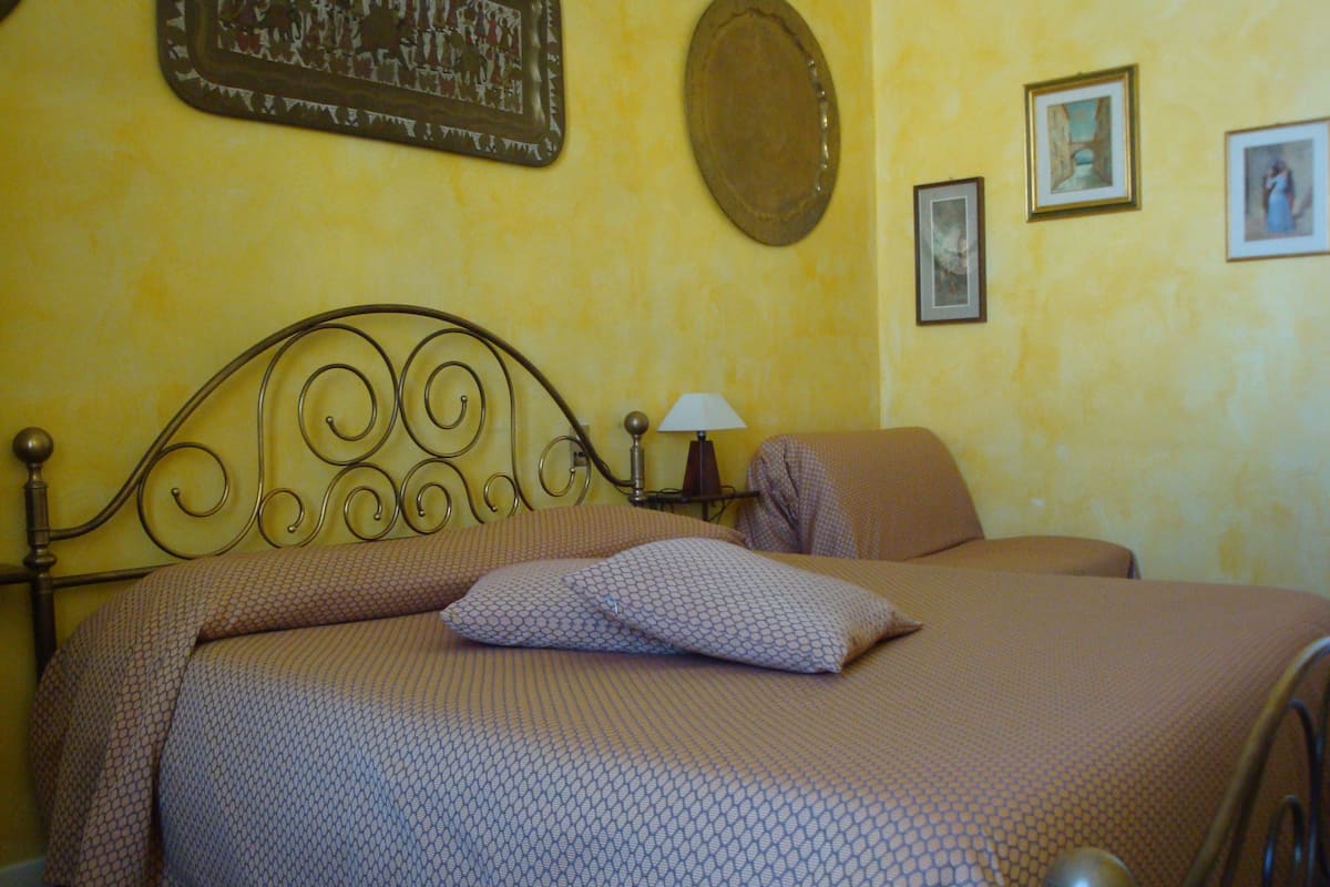A bed with a metal headboard, a patterned bedspread, and pillows. There is a wall-mounted lamp and framed pictures on the wall.