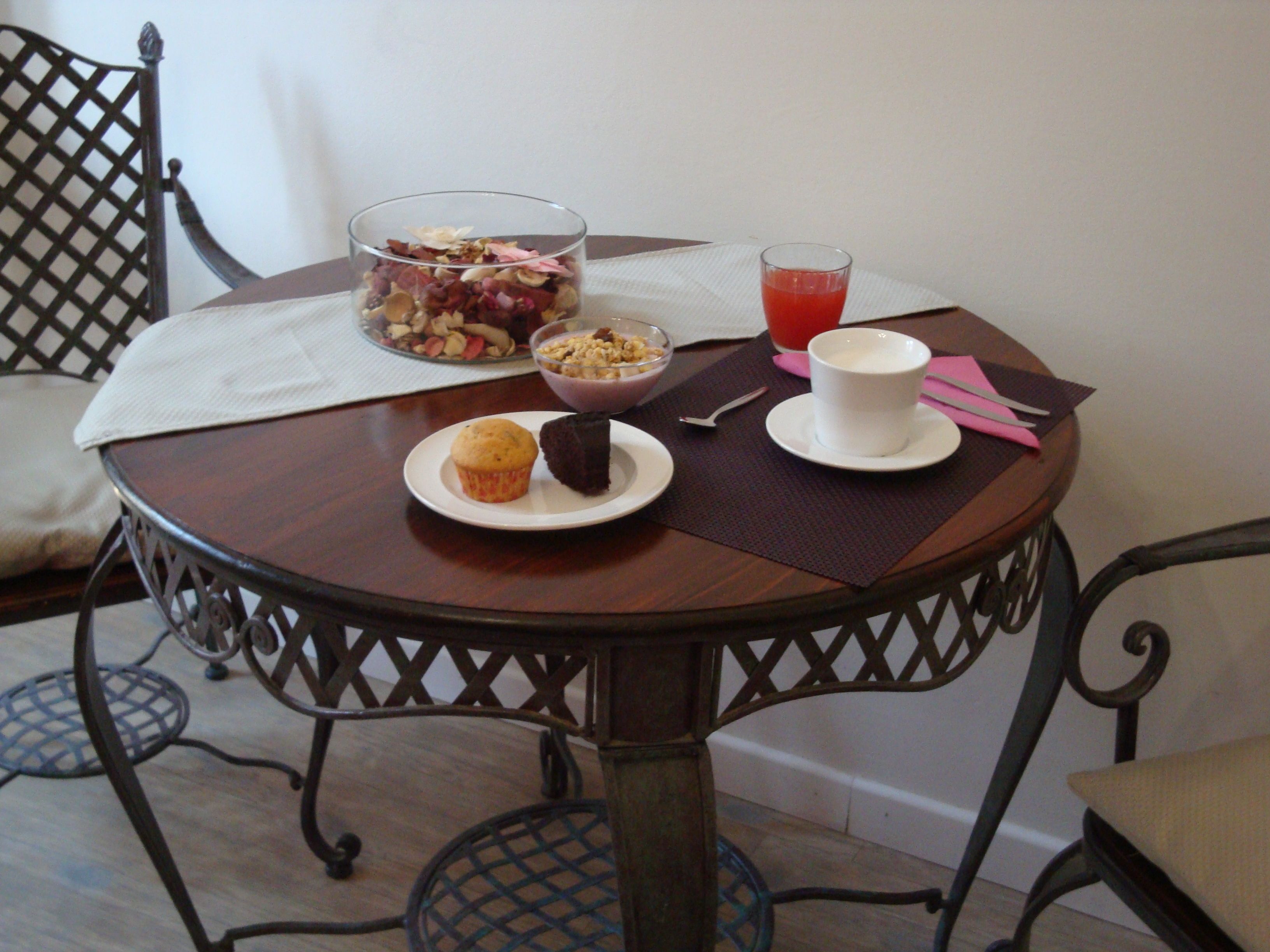 A round dining table with a white placemat, a bowl of cereal, a glass of red juice, a muffin, and a cup of coffee.