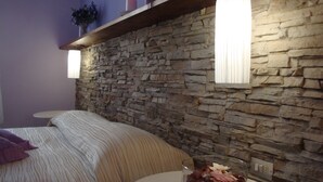 Un dormitorio con pared de piedra, una cama con ropa de cama a rayas, una mesita redonda con un cuenco de cristal con flores secas y una estantería con velas.