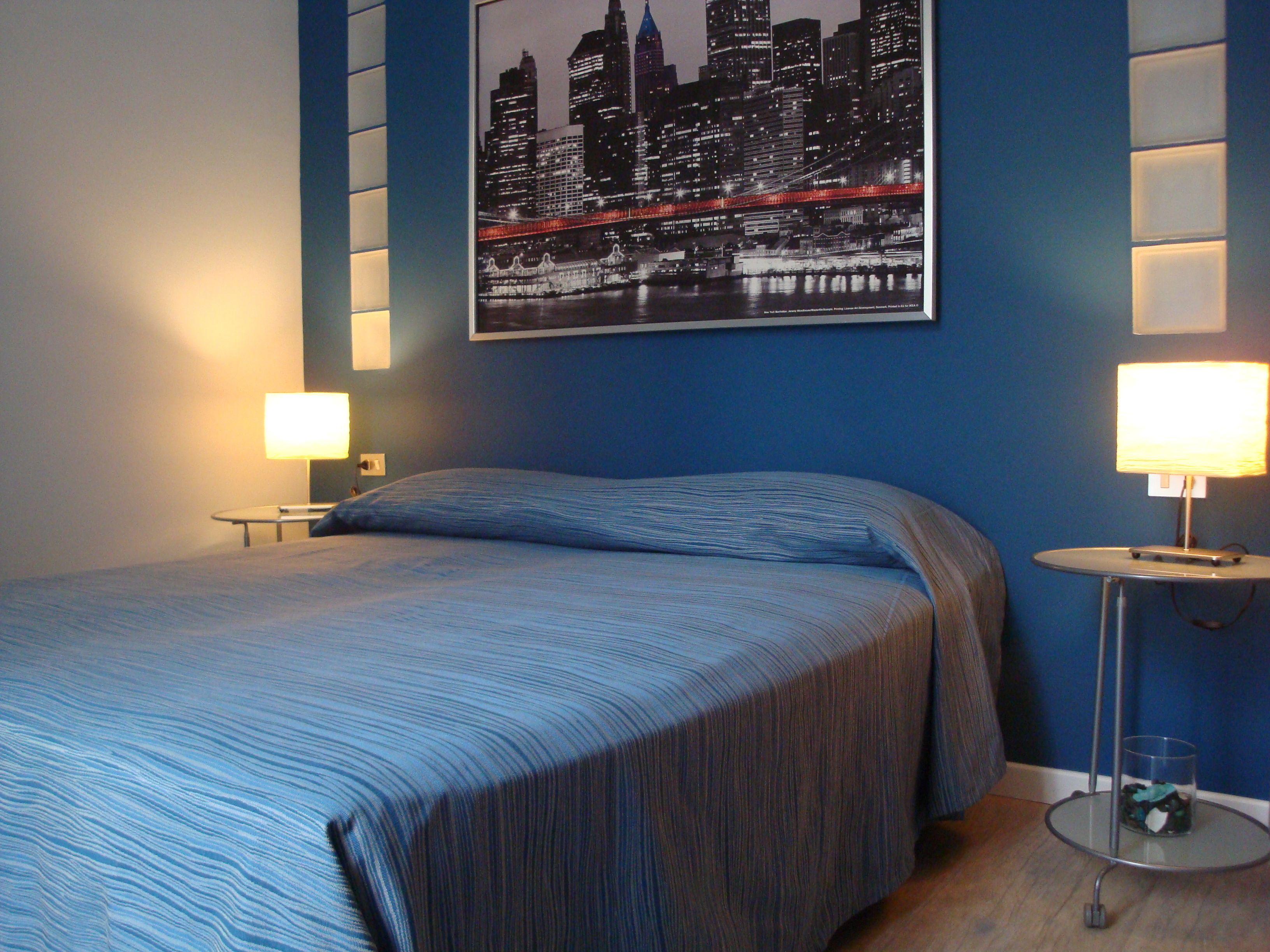 A hotel room with a bed, two bedside lamps, a framed cityscape picture on the wall, and a small table with a glass.